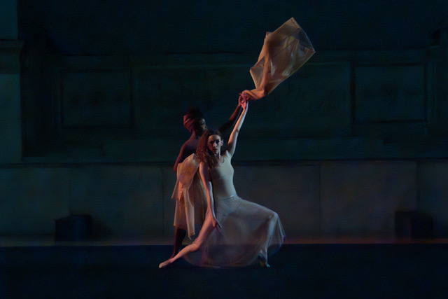 Photo of Noi-Tar-Gim by Kevin Iega Jeff. Performed by New York Theatre Ballet. Two women are against a dark background. One woman, (white, fair-skinned, long curly hair to the right side) faces forward with legs bent into a lunge position. Her left are is raised into the air as she throws a jacket held in her left hand. The other woman (black, medium brown-skinned, hair wrapped up in a scarf above her head), stands behind the first women and slightly to her right. She faces left as she holds other jackets in her arms.